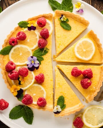 Lemon tart with raspberries, lemon slices, mint leaves, and edible flowers on top, placed on a white plate.