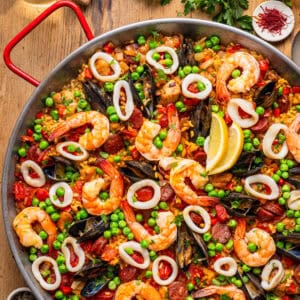 Colorful seafood paella in a pan with shrimp, mussels, calamari rings, peas, chorizo, rice, tomato, and lemon wedges.