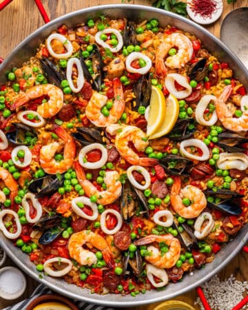 A large pan of seafood paella with shrimp, mussels, squid rings, peas, red peppers, and lemon wedges.