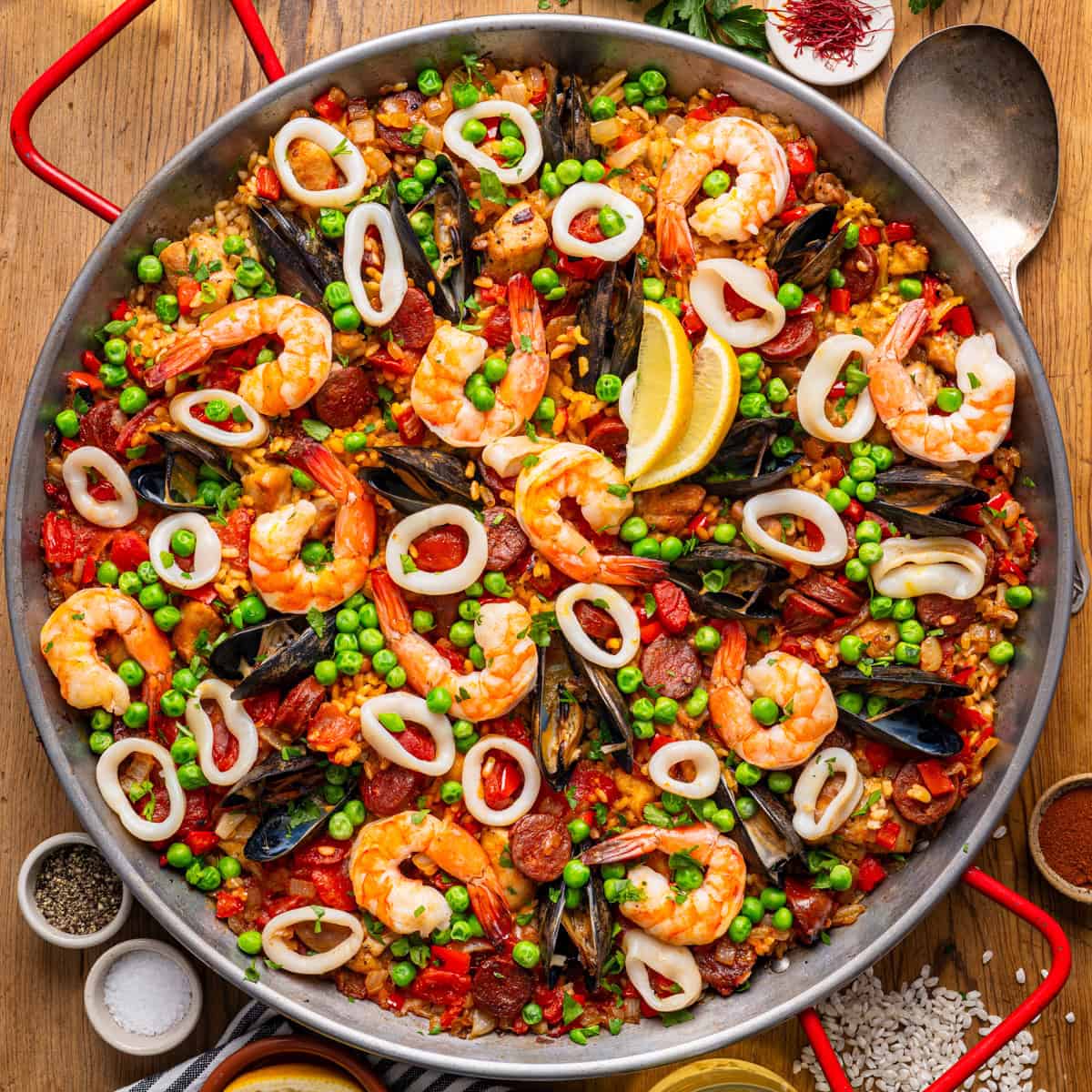A large pan of seafood paella with shrimp, mussels, squid rings, peas, red peppers, and lemon wedges.