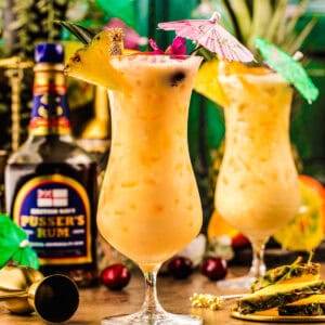 Two tropical cocktails in hurricane glasses with fruit garnishes and paper umbrellas.