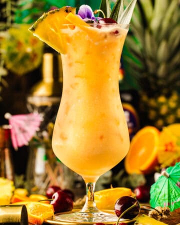 A tropical cocktail in a curved glass, garnished with a pineapple slice, cherry, and edible flower.