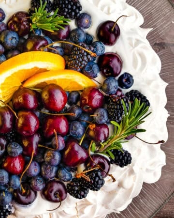A pavlova topped with whipped cream, cherries, blueberries, blackberries, orange slices, and rosemary.
