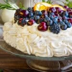 A pavlova dessert topped with blueberries, blackberries, cherries, and orange slices, garnished with rosemary.