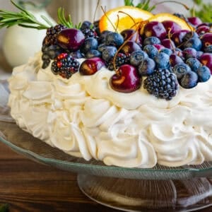 A pavlova dessert topped with blueberries, blackberries, cherries, and orange slices, garnished with rosemary.