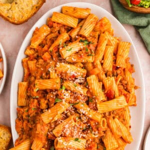 A white platter filled with rigatoni pasta in a rich tomato sauce, garnished with grated cheese and parsley.