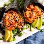 Two hand-rolled sushi cones with spicy tuna, avocado slices, rice, and microgreens, wrapped in nori and topped with black sesame seeds, on a white plate.