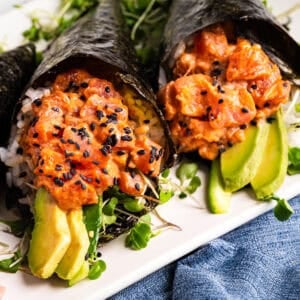 Two hand-rolled sushi cones with spicy tuna, avocado slices, rice, and microgreens, wrapped in nori and topped with black sesame seeds, on a white plate.