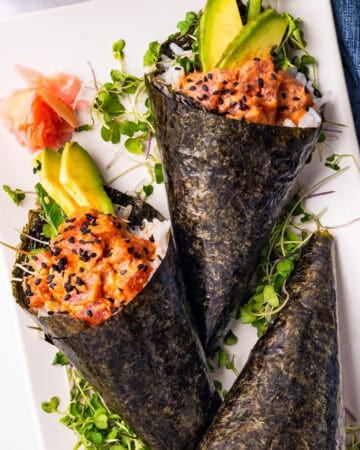 Two hand-rolled sushi cones filled with spicy salmon, avocado, rice, and microgreens, wrapped in seaweed, garnished with black sesame seeds and served with pickled ginger on a white plate.