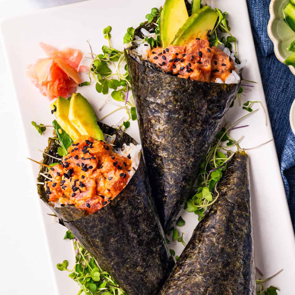 Two hand-rolled sushi cones filled with spicy salmon, avocado, rice, and microgreens, wrapped in seaweed, garnished with black sesame seeds and served with pickled ginger on a white plate.
