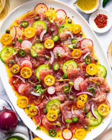 A white oval platter with thin slices of raw fish, cucumber, radish, kumquat, capers, red onion, chili flakes, and dressing.