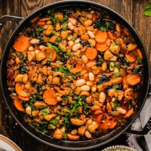 A pot of hearty vegetable stew with carrots, white beans, greens, and chopped herbs in a rich broth, sitting on a rustic wooden table.