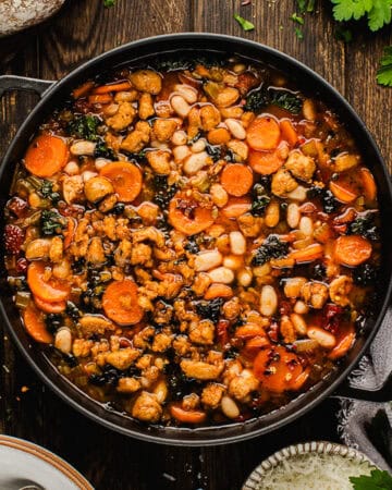 A pot of hearty vegetable stew with carrots, white beans, leafy greens, and crumbled sausage, garnished with herbs, sits on a wooden table.