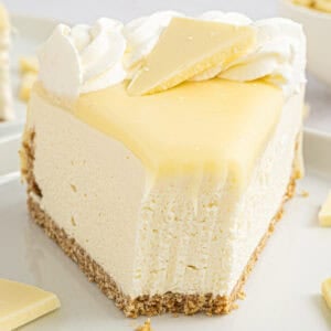 A slice of creamy white cheesecake with a graham cracker crust, topped with whipped cream and a piece of white chocolate, sits on a plate.