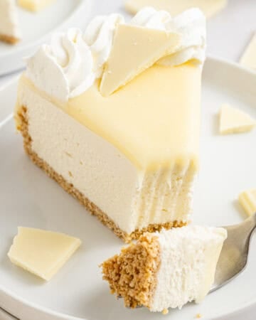 A slice of white chocolate cheesecake with a graham cracker crust, topped with whipped cream and white chocolate pieces.