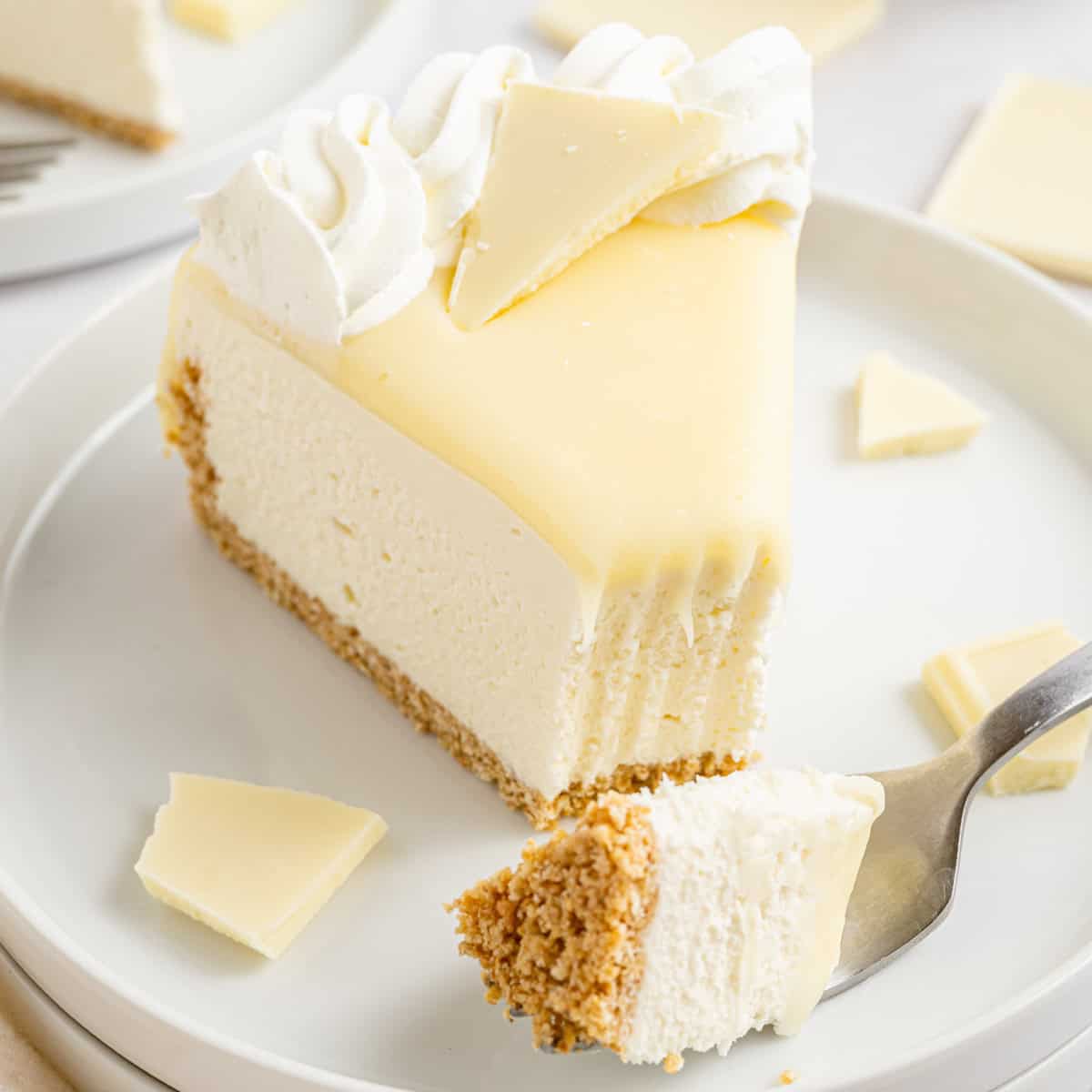 A slice of white chocolate cheesecake with a graham cracker crust, topped with whipped cream and white chocolate pieces.