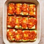A baking dish filled with zucchini roll-ups topped with chunky red tomato sauce, arranged neatly in rows and ready to be baked.