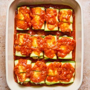 A baking dish filled with zucchini roll-ups topped with chunky red tomato sauce, arranged neatly in rows and ready to be baked.
