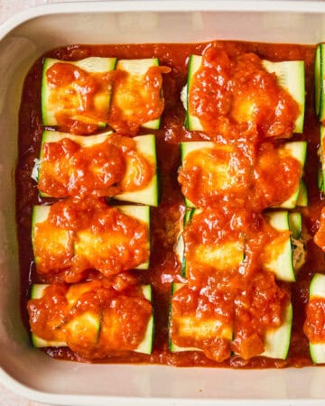 A baking dish filled with zucchini roll-ups topped with tomato sauce, arranged in neat rows and ready to be baked.
