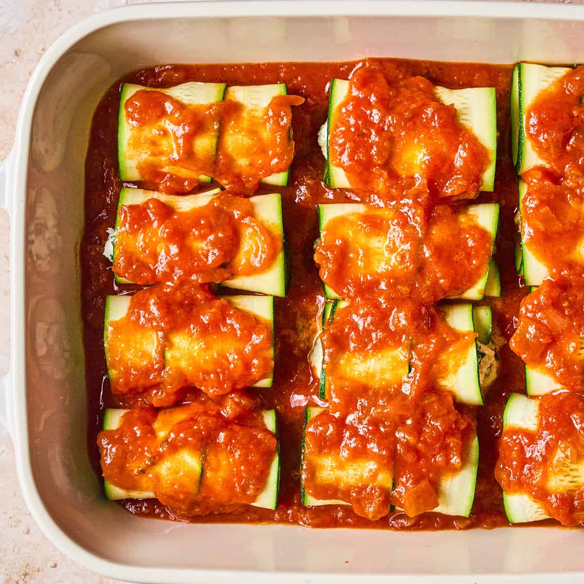 A baking dish filled with zucchini roll-ups topped with tomato sauce, arranged in neat rows and ready to be baked.