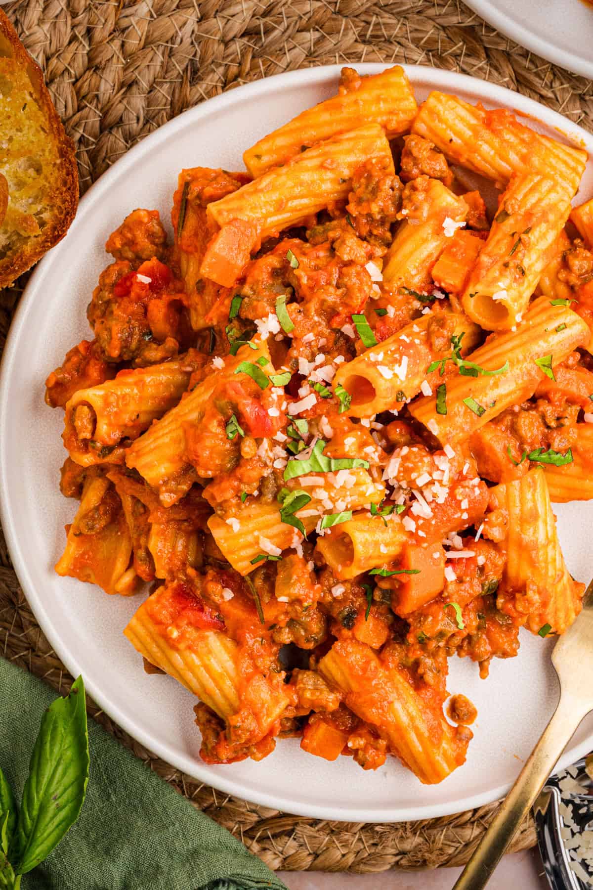 Sausage Ragu Plate of rigatoni pasta with meat sauce, sprinkled with grated cheese and chopped herbs, on a woven placemat.
