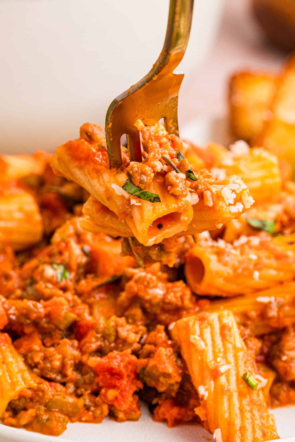 Sausage Ragu A fork lifts rigatoni pasta topped with meat sauce and grated cheese above a plate filled with the same pasta dish.