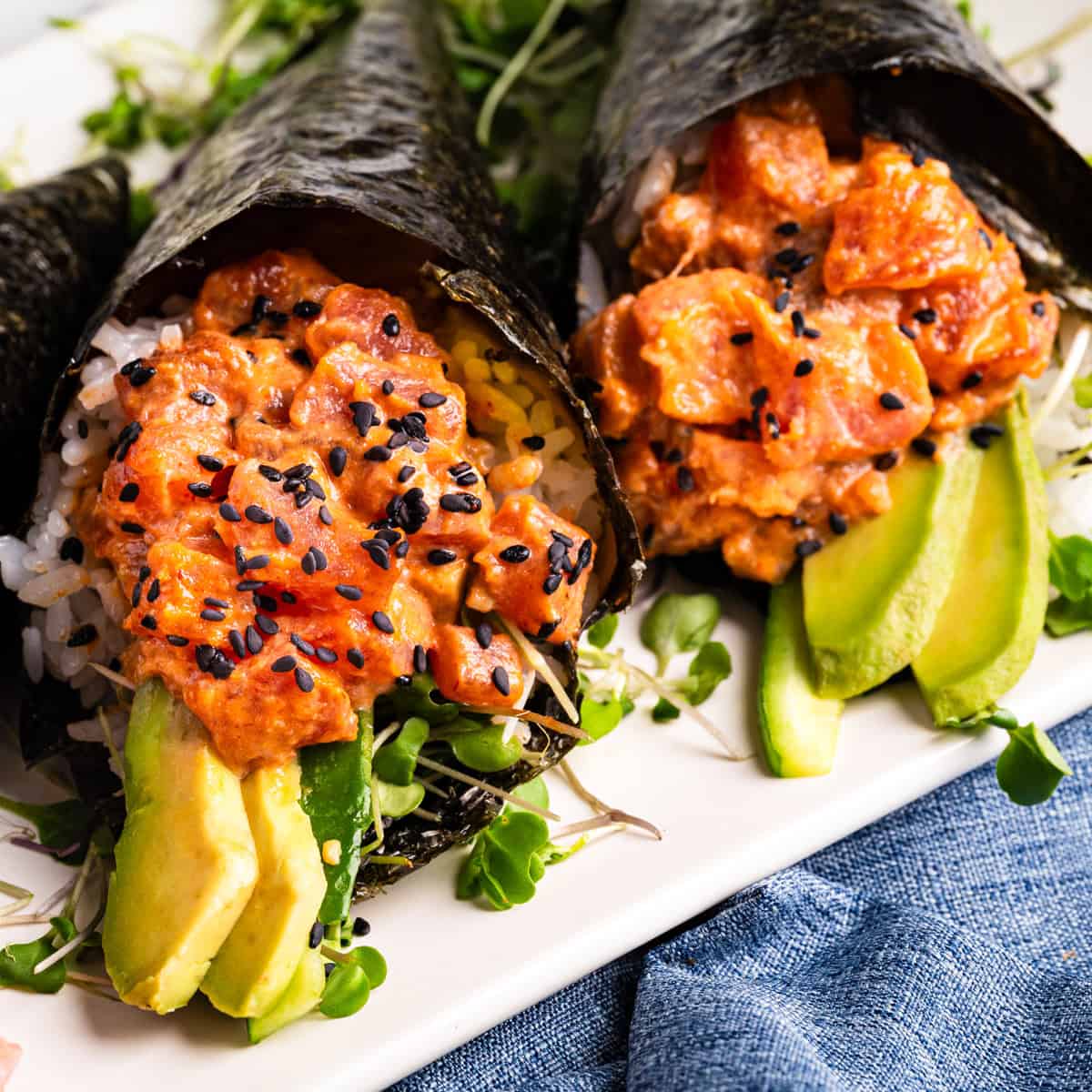 Two hand-rolled sushi cones filled with spicy diced fish, sliced avocado, rice, and black sesame seeds, wrapped in seaweed, garnished with sprouts on a white plate.