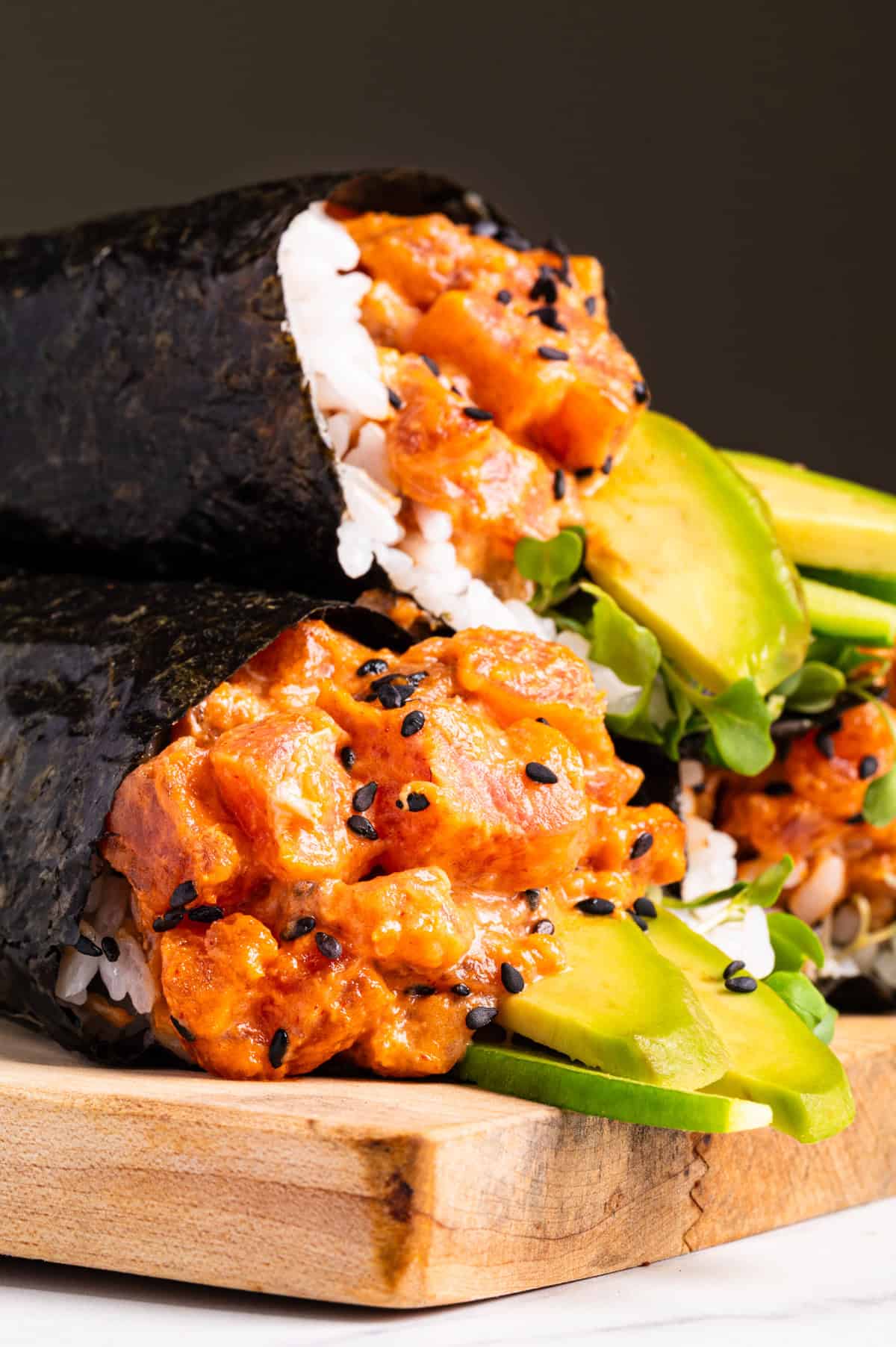 Spicy Tuna Hand Rolls Two sushi burritos filled with spicy tuna, rice, avocado slices, greens, and black sesame seeds, wrapped in seaweed, stacked on a wooden board.