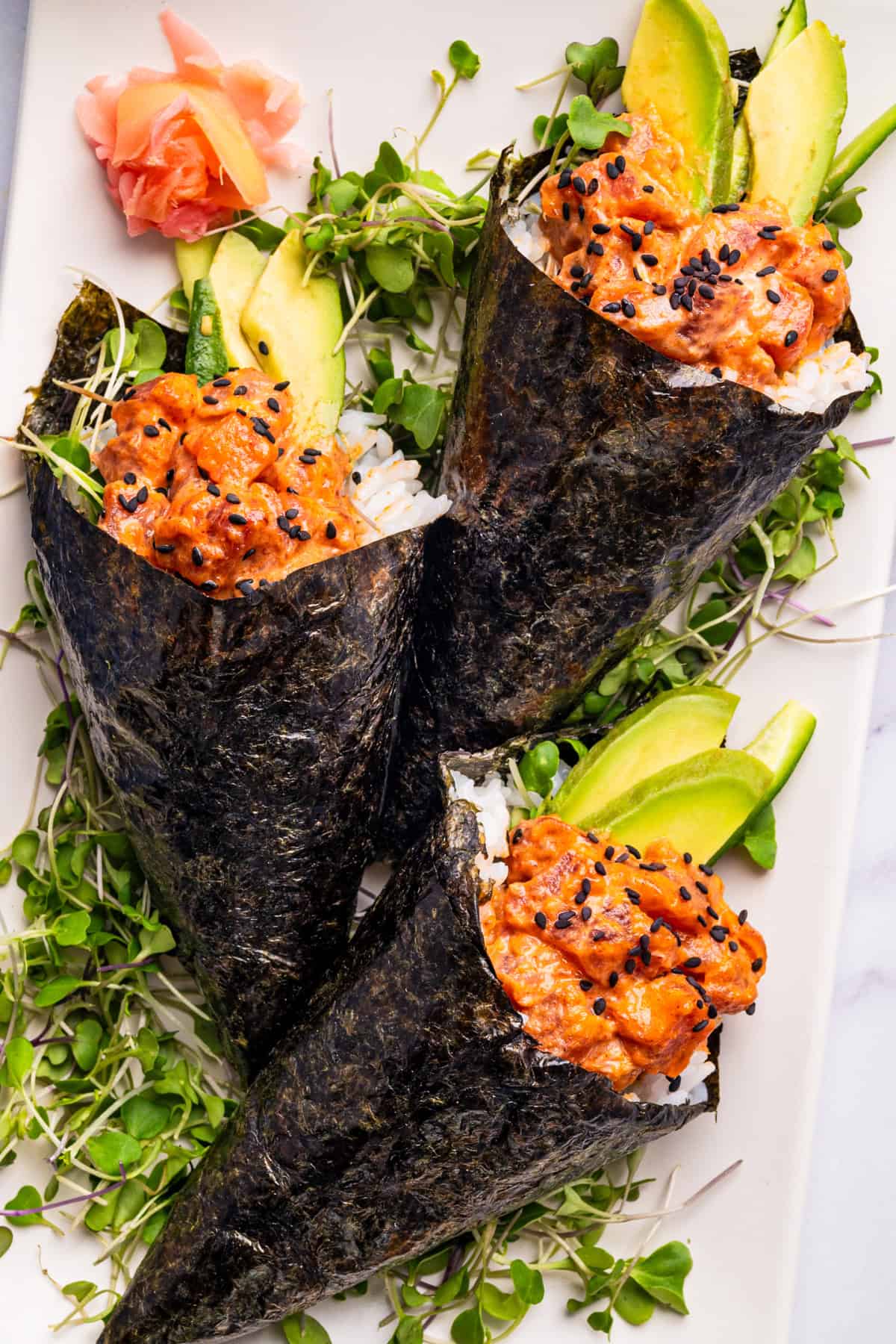 Spicy Tuna Hand Rolls Three sushi rolls filled with avocado slices, rice, and black sesame seeds, wrapped in seaweed, garnished with microgreens.