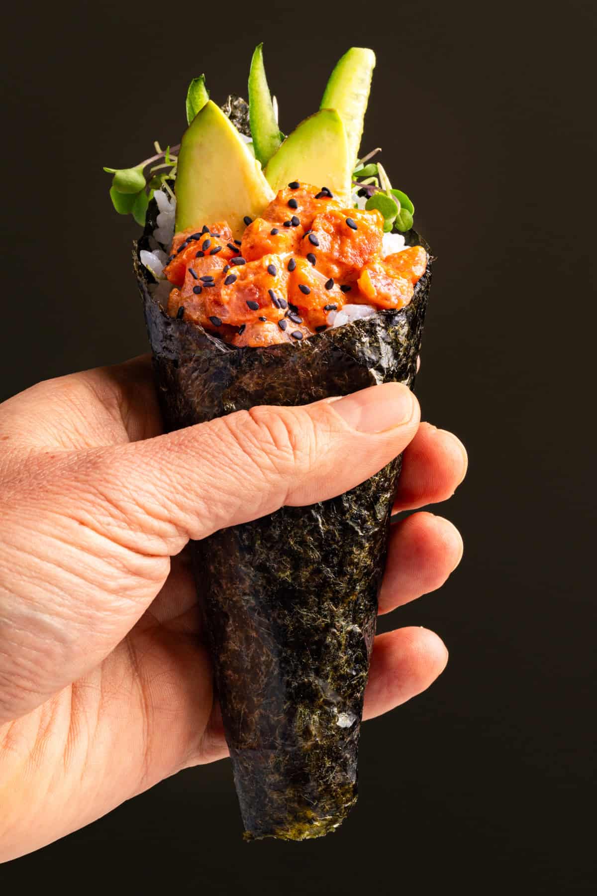 Spicy Tuna Hand Rolls A hand holding a sushi hand roll with seaweed wrap, rice, spicy diced fish, black sesame seeds, avocado slices, and microgreens against a plain dark background.