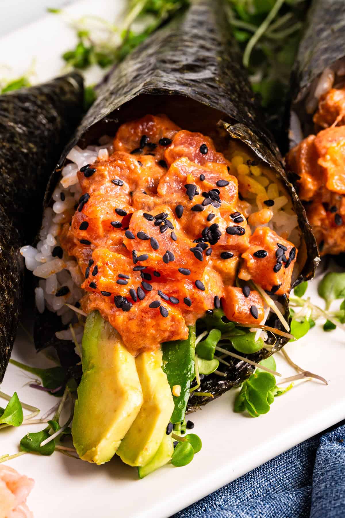 Spicy Tuna Hand Rolls Close-up of a sushi hand roll filled with rice, avocado slices, spicy tuna mix, and sprinkled with black sesame seeds, wrapped in nori seaweed, and garnished with microgreens.