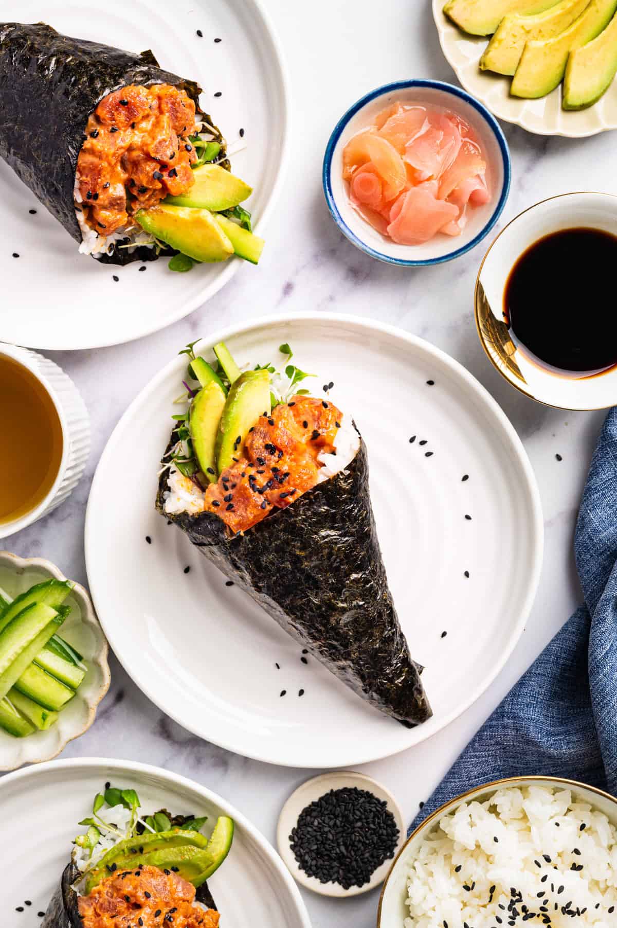 Spicy Tuna Hand Rolls Two sushi hand rolls filled with spicy tuna, avocado slices, and sprouts are on white plates.