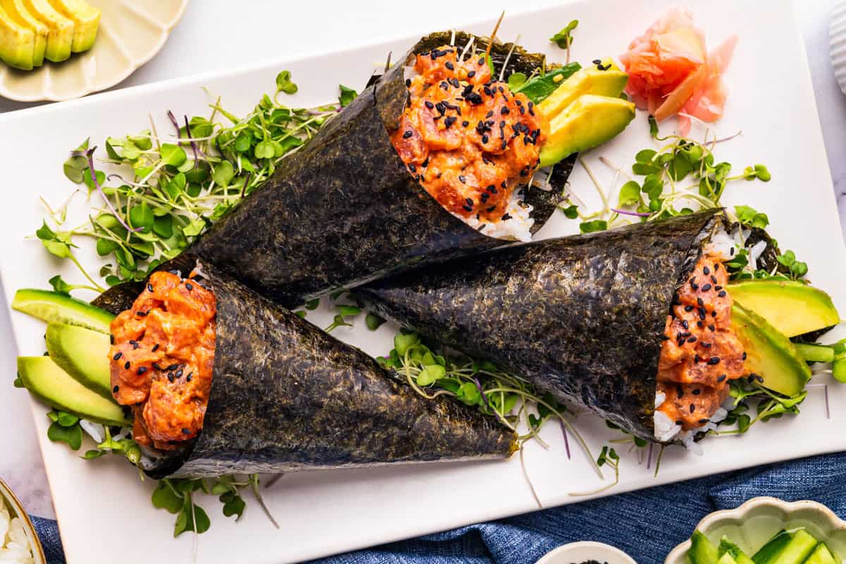 Spicy Tuna Hand Rolls Three spicy tuna hand rolls with avocado, rice, microgreens, and black sesame seeds are arranged on a white platter.