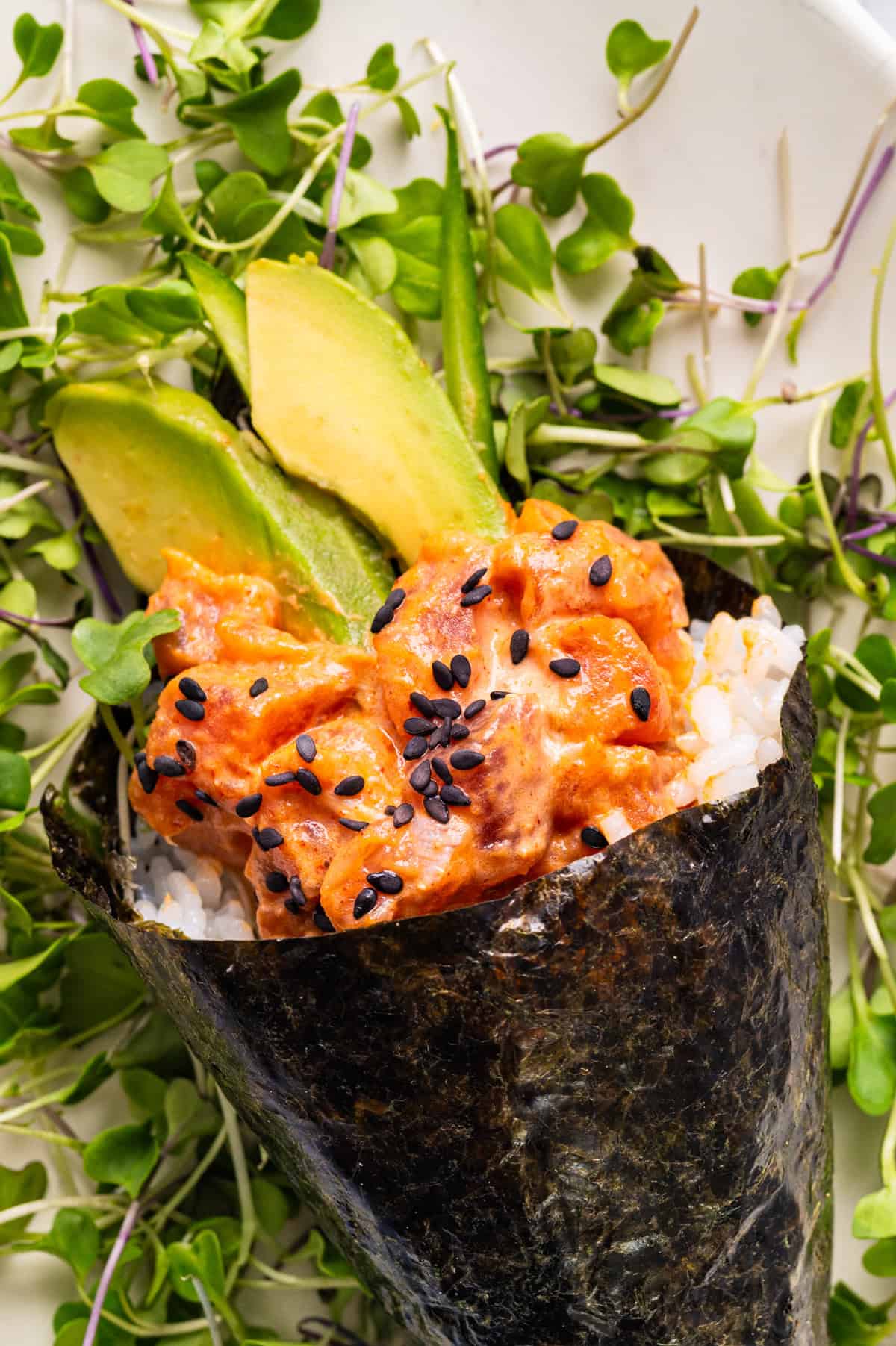 Spicy Tuna Hand Rolls A hand roll sushi with spicy tuna, avocado slices, white rice, and black sesame seeds, wrapped in seaweed, garnished with fresh microgreens on a white plate.