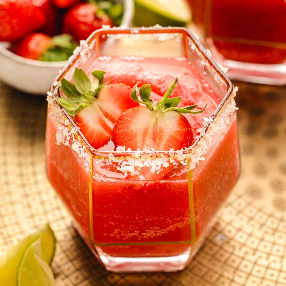 Strawberry Margarita A hexagonal glass of bright red strawberry drink with ice, salt-rimmed with salt, and garnished with halved strawberries.