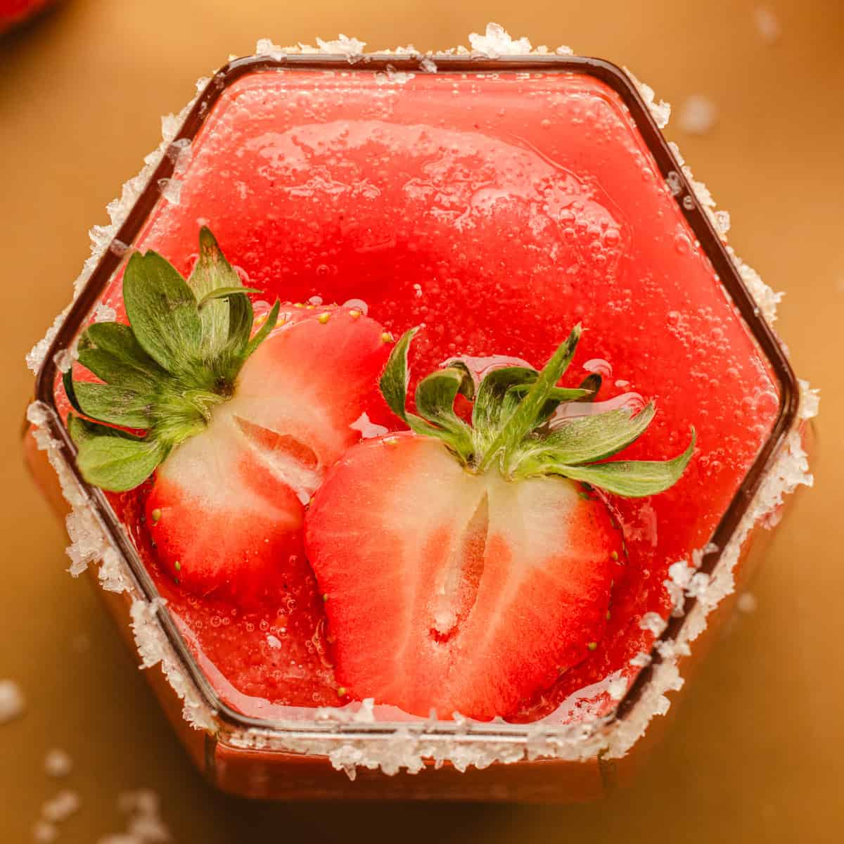 Strawberry Margarita A hexagonal glass filled with a bright red drink, topped with two fresh strawberry halves and a salted rim.