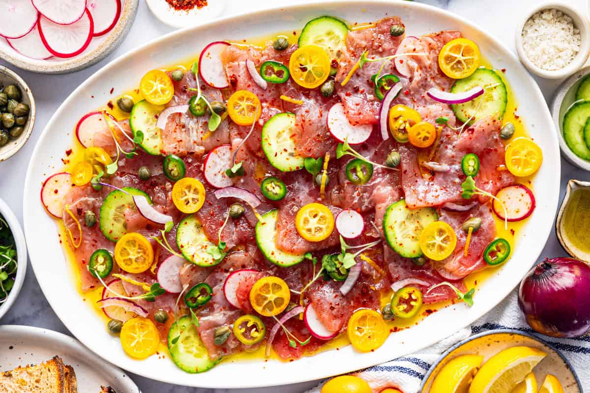 Oval platter of thinly sliced raw fish topped with cucumbers, radishes, kumquat slices, capers, red onion, herbs, and drizzled oil.