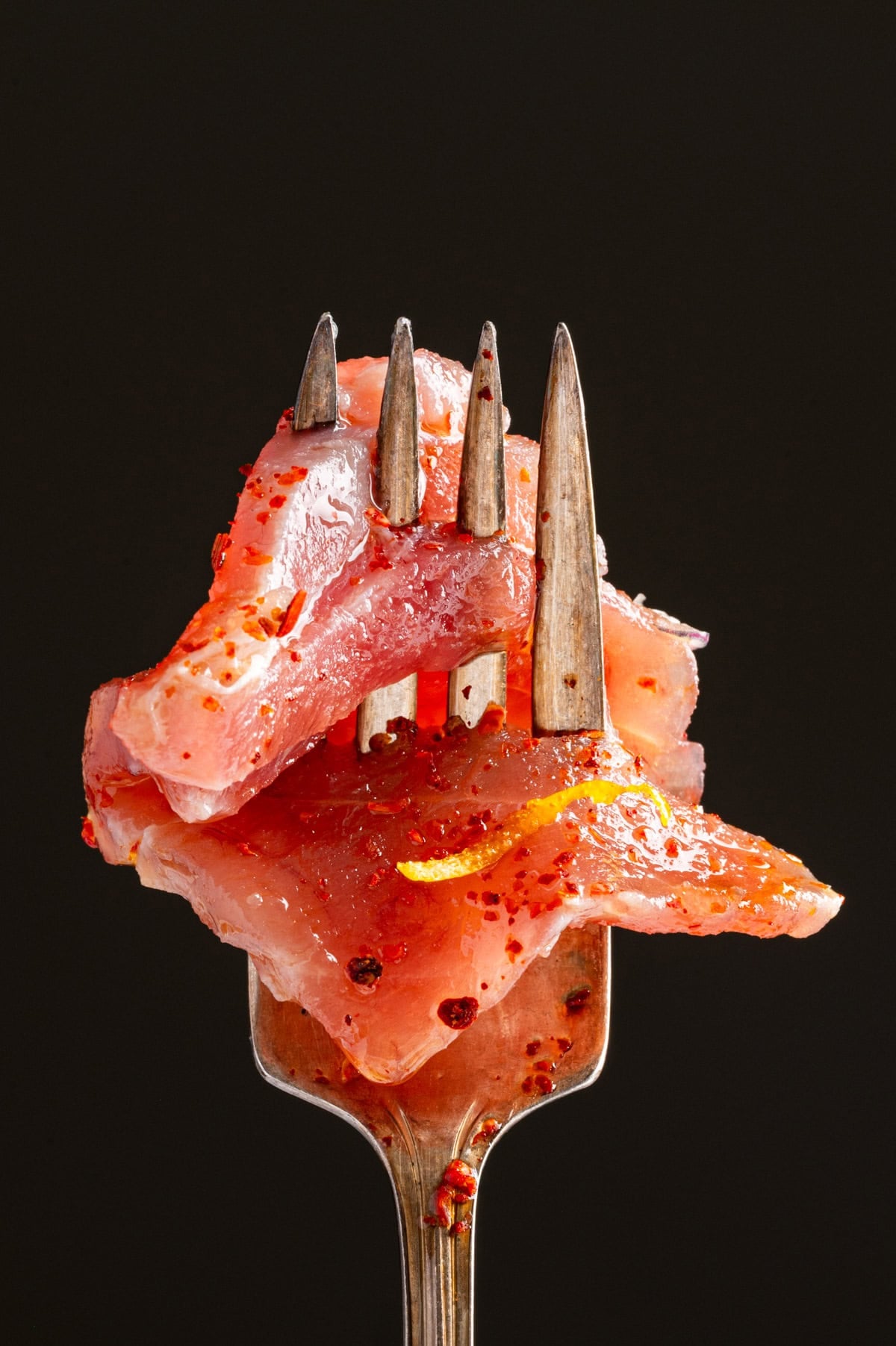 Tuna Crudo A close-up of raw, seasoned fish slices on a fork. The fish is sprinkled with red spices and garnished with a small piece of lemon zest.