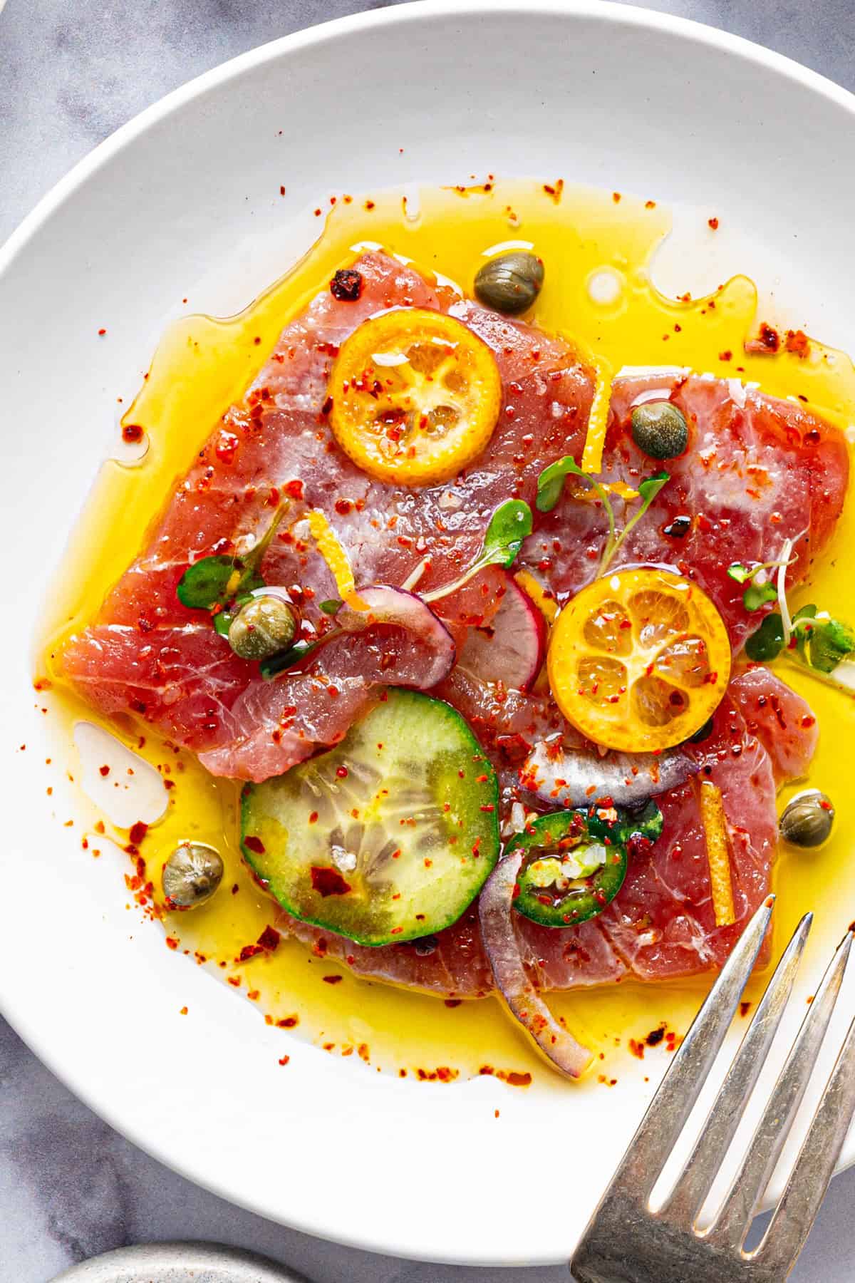 Tuna Crudo A white plate with slices of raw fish, cucumber, capers, microgreens, red onion, lemon slices, chili flakes, and drizzled olive oil.