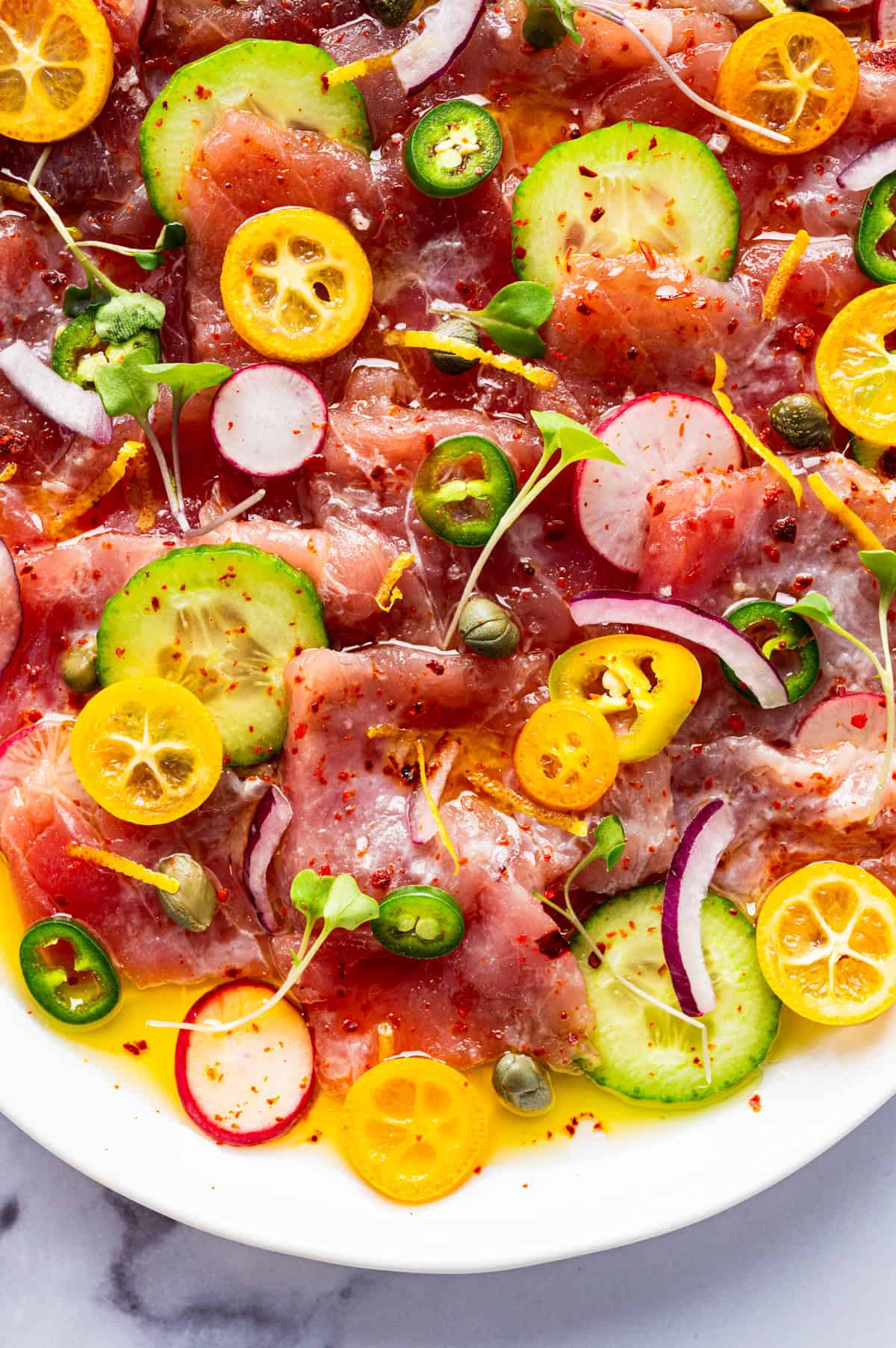 Tuna Crudo Colorful plate of tuna topped with sliced cucumber, radish, kumquat, red onion, capers, microgreens, and red pepper flakes, drizzled with oil.