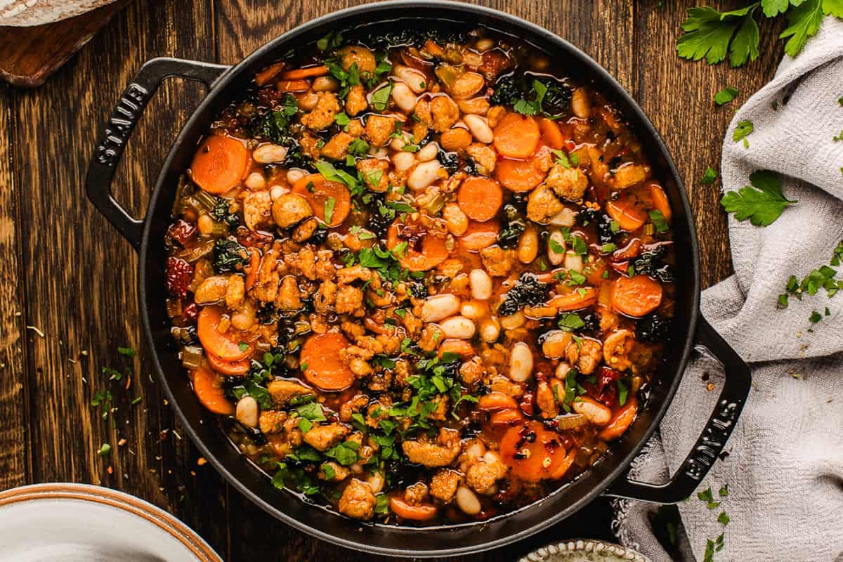 Tuscan White Bean Soup A black pot holds a colorful Tuscan White Bean Soup with white beans, chopped carrots, leafy greens, and crumbled sausage, garnished with fresh herbs.