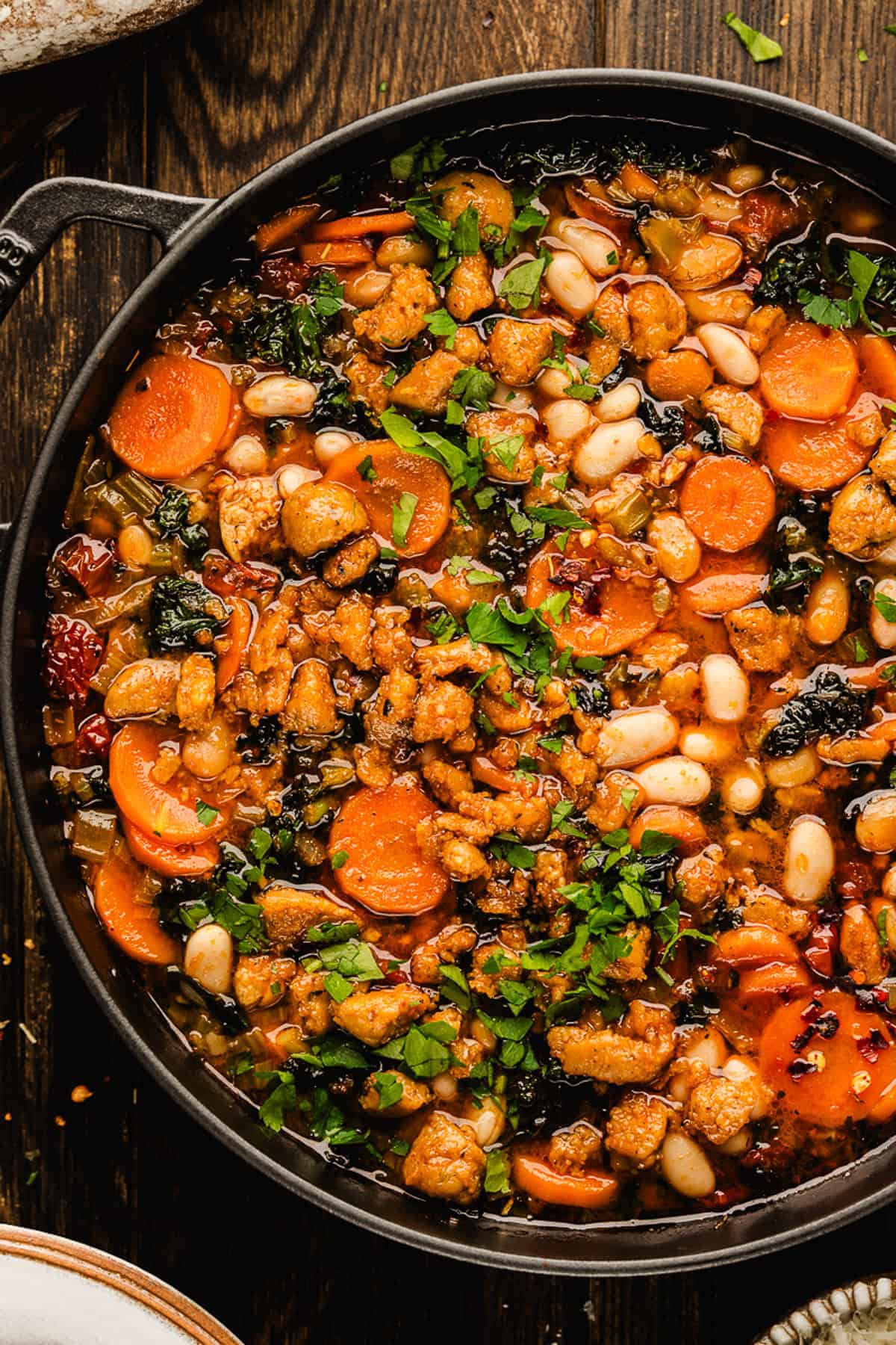 Tuscan White Bean Soup A close-up of Tuscan White Bean Soup in a black pot, filled with white beans, sliced carrots, greens, and crumbled sausage, topped with chopped parsley.
