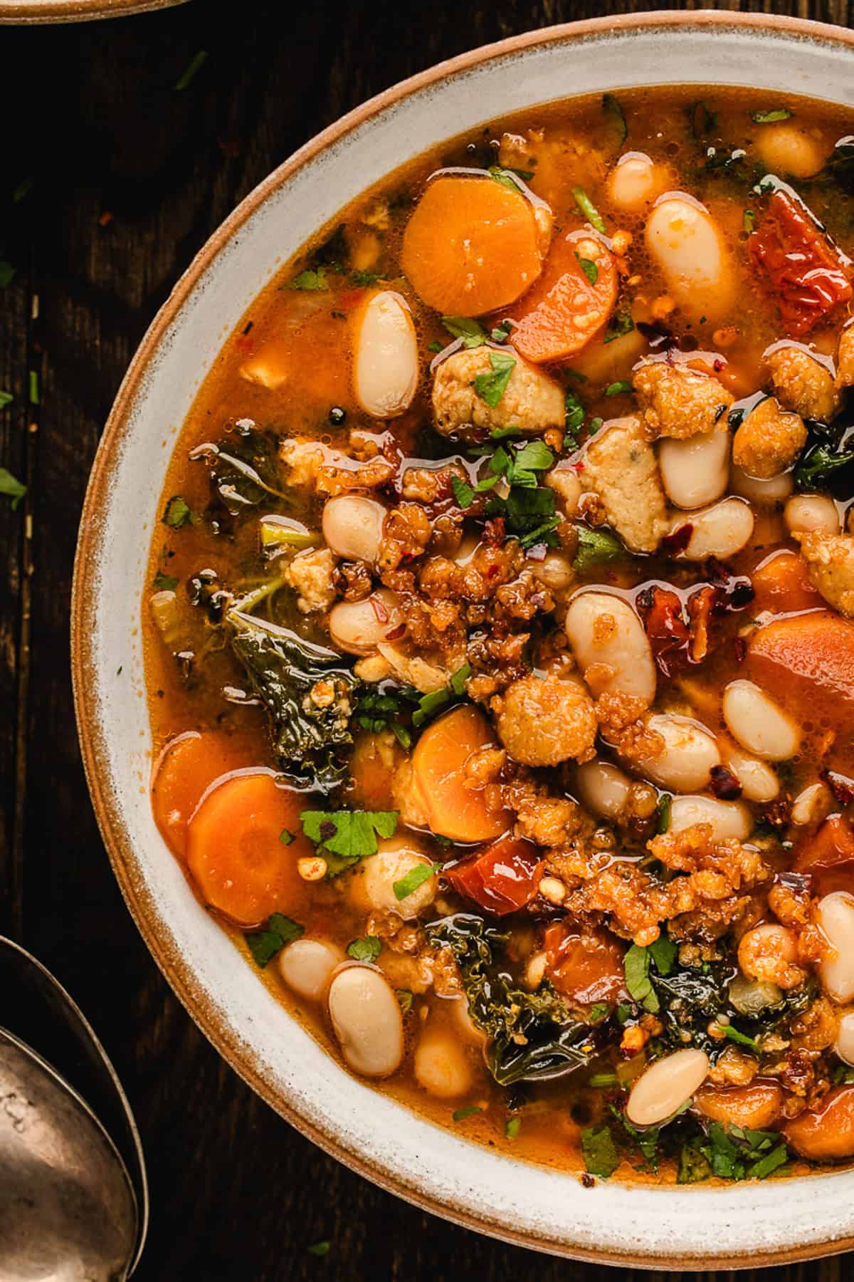 Tuscan White Bean Soup A bowl of hearty Tuscan White Bean Soup with white beans, carrots, kale, tomatoes, crumbled sausage, and fresh herbs in a rich broth, viewed from above.