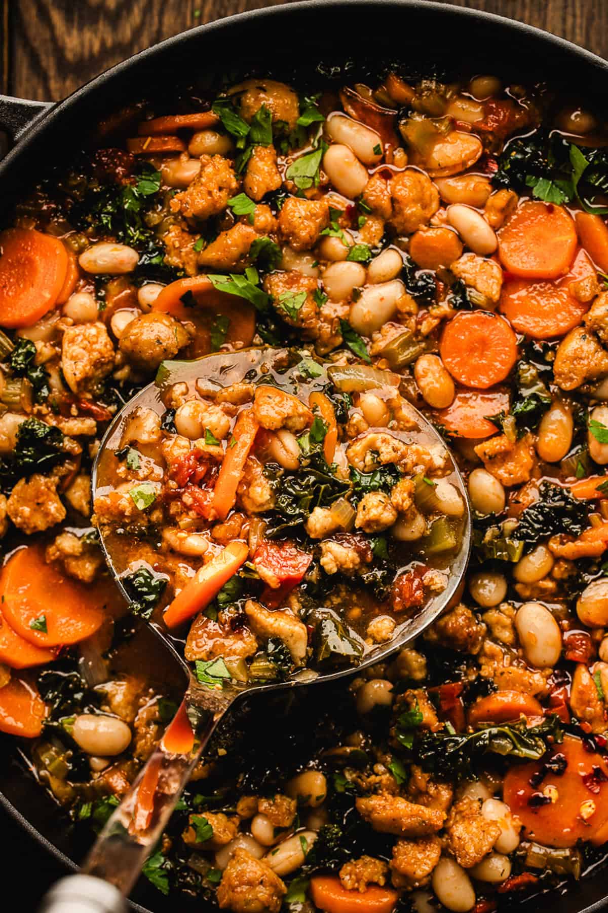 Tuscan White Bean Soup A ladle scoops up Tuscan White Bean Soup with white beans, carrots, leafy greens, ground meat, and herbs from a pot.