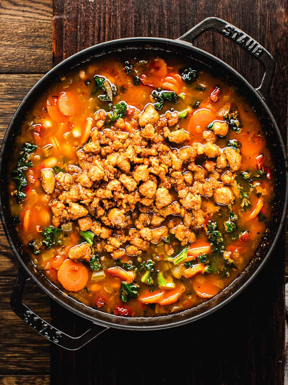 Tuscan White Bean Soup A pot of hearty Tuscan White Bean Soup with sliced carrots, beans, kale, and other vegetables, topped with crumbled browned sausage, on a wooden surface.