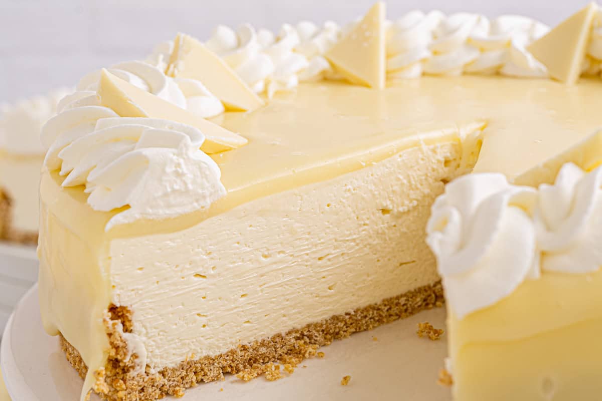 A creamy white cheesecake topped with whipped cream and white chocolate triangles sits on a crust, with one large slice removed to show the smooth texture inside.