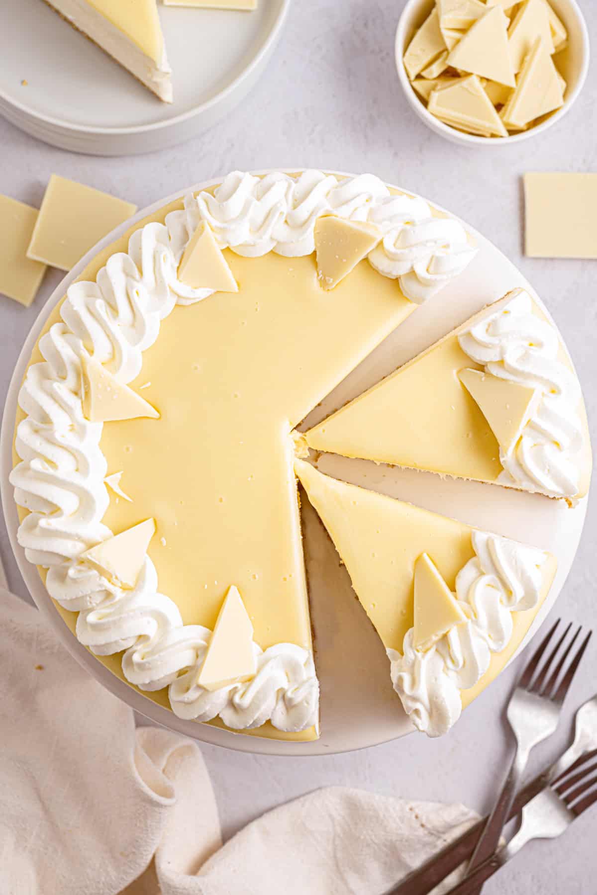 White Chocolate Cheesecake A round white chocolate cheesecake topped with whipped cream swirls and white chocolate triangles, with two slices cut.