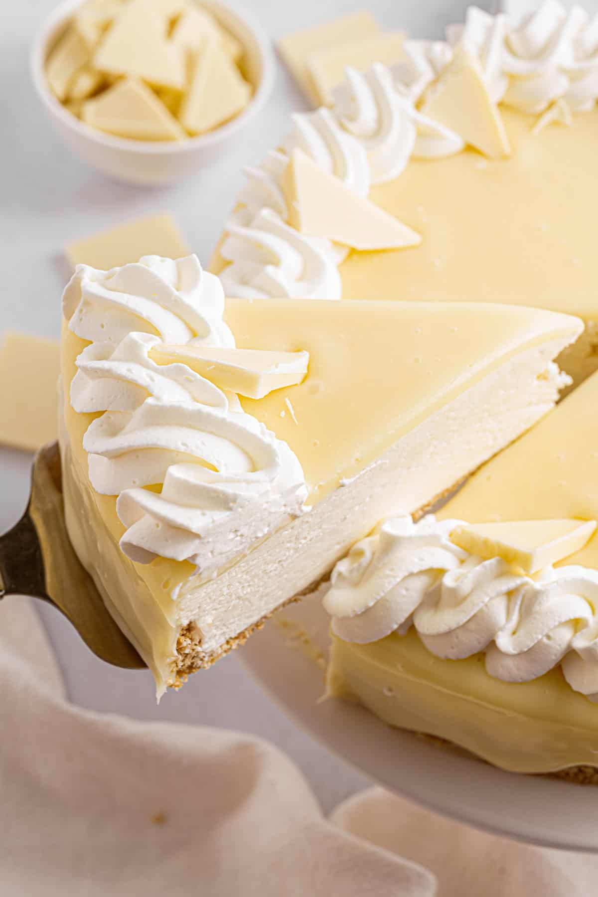 White Chocolate Cheesecake A slice of creamy white chocolate cheesecake with whipped cream is being lifted from a whole cheesecake, which is also decorated with white chocolate pieces.