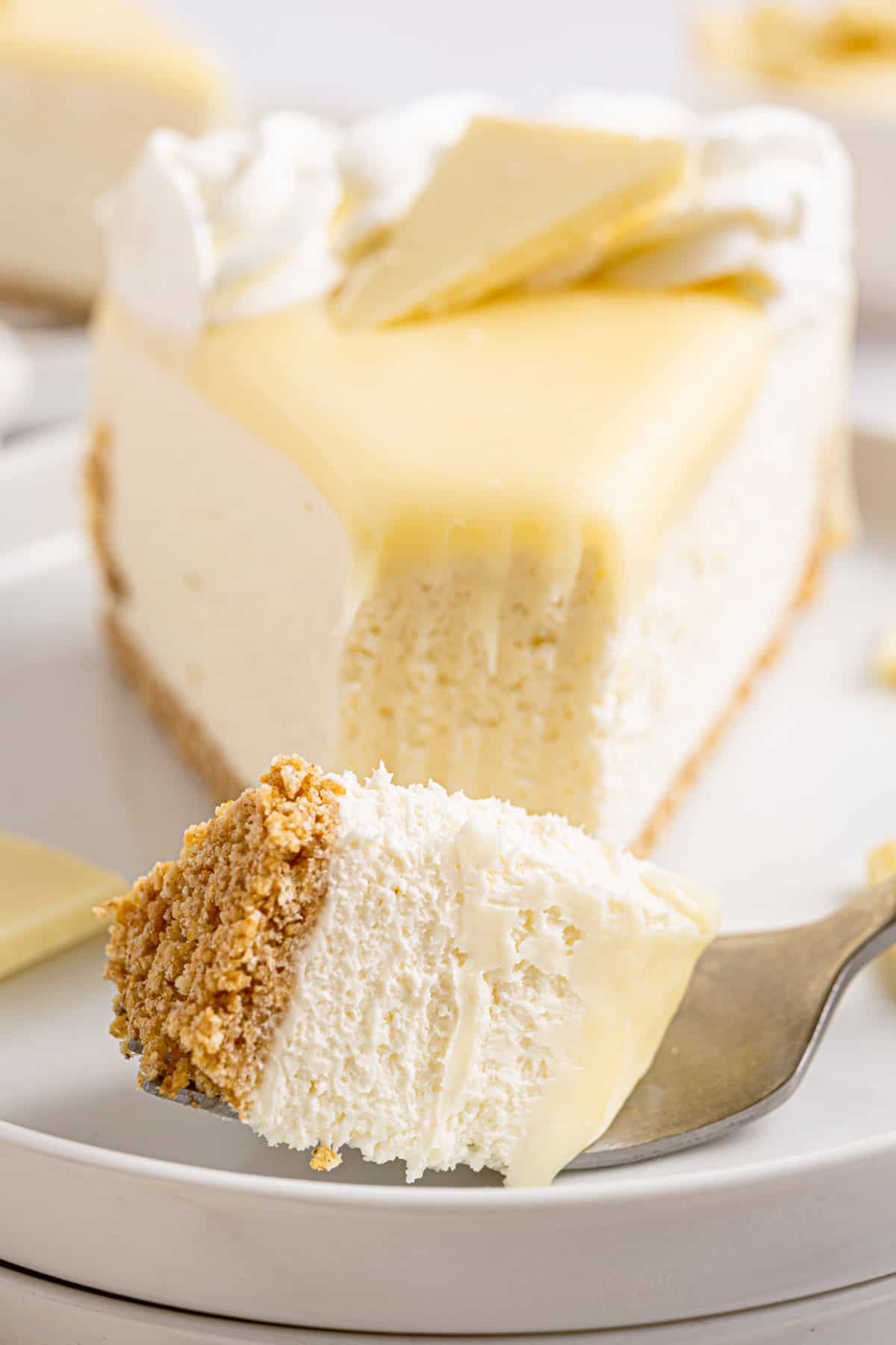 White Chocolate Cheesecake A close-up of a creamy cheesecake slice with a graham cracker crust, topped with whipped cream and white chocolate.