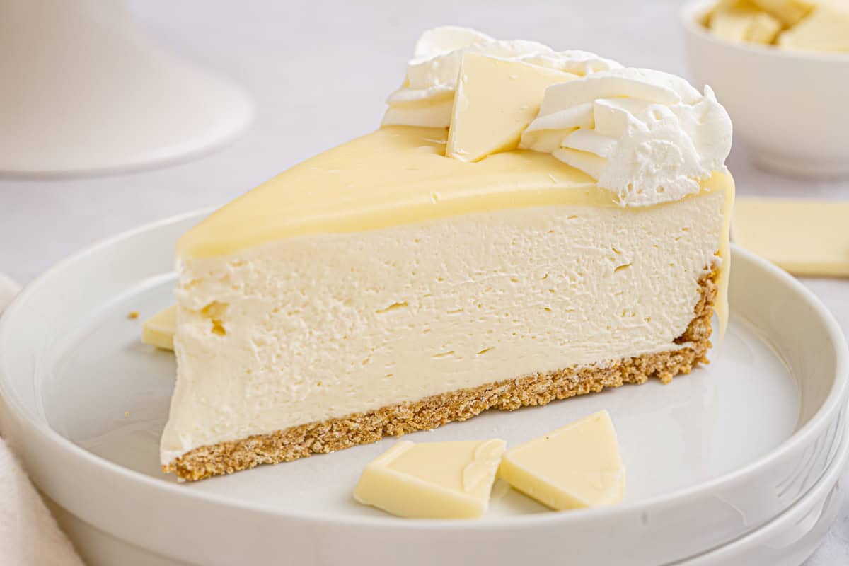 White Chocolate Cheesecake A slice of creamy white cheesecake with a graham cracker crust, topped with whipped cream and white chocolate pieces.
